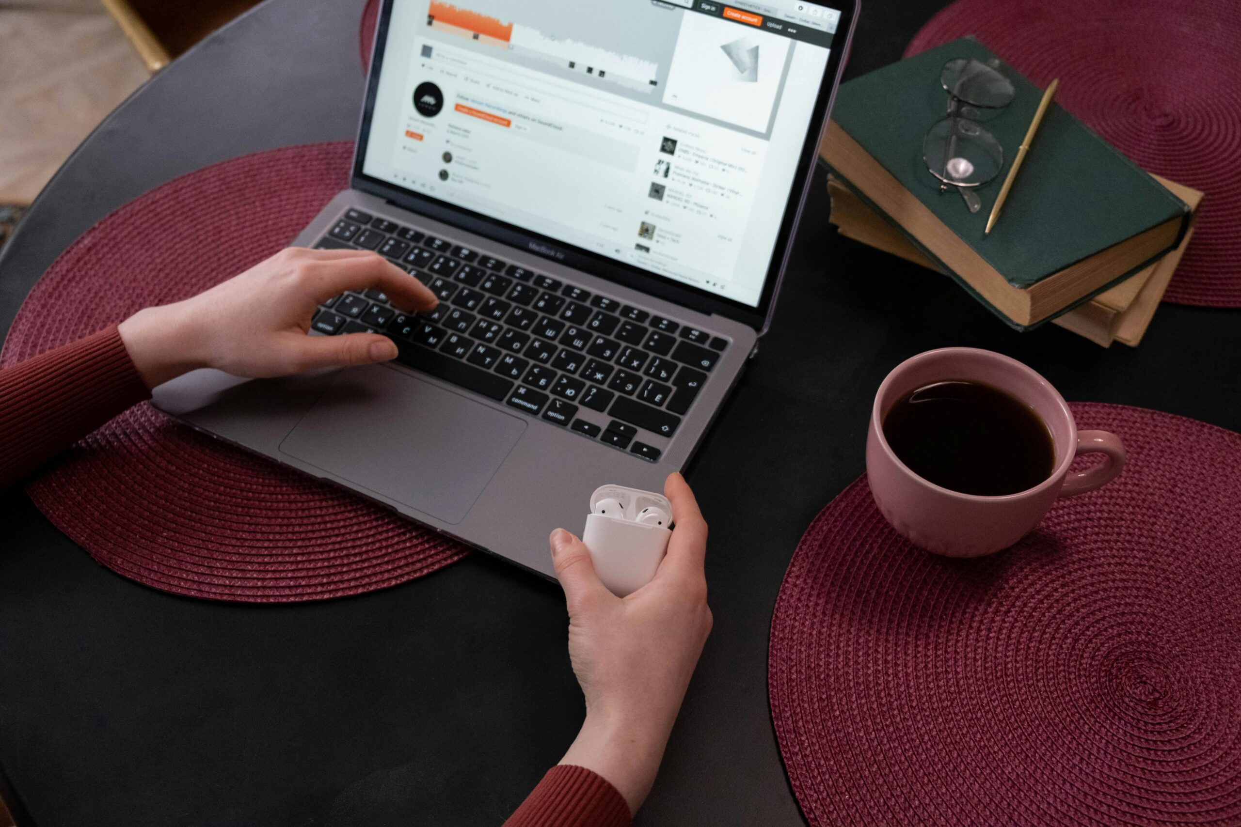 Close-up of hands typing on a laptop with a cup of tea, books, and wireless earbuds.