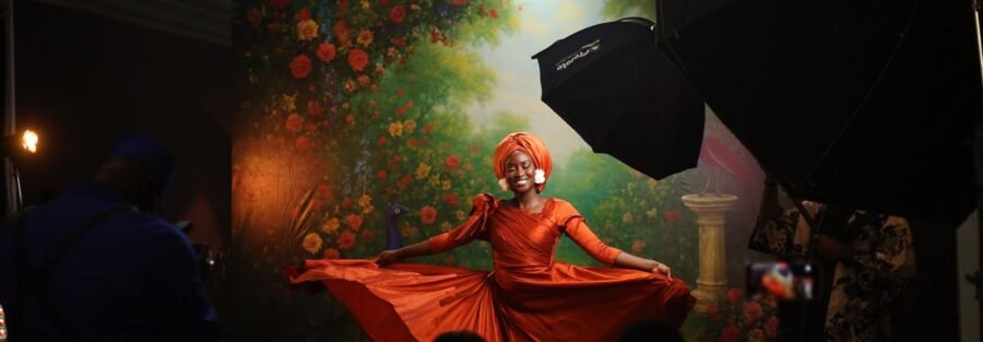 A woman in an orange dress poses gracefully indoors with a vibrant floral backdrop in Abuja, Nigeria.