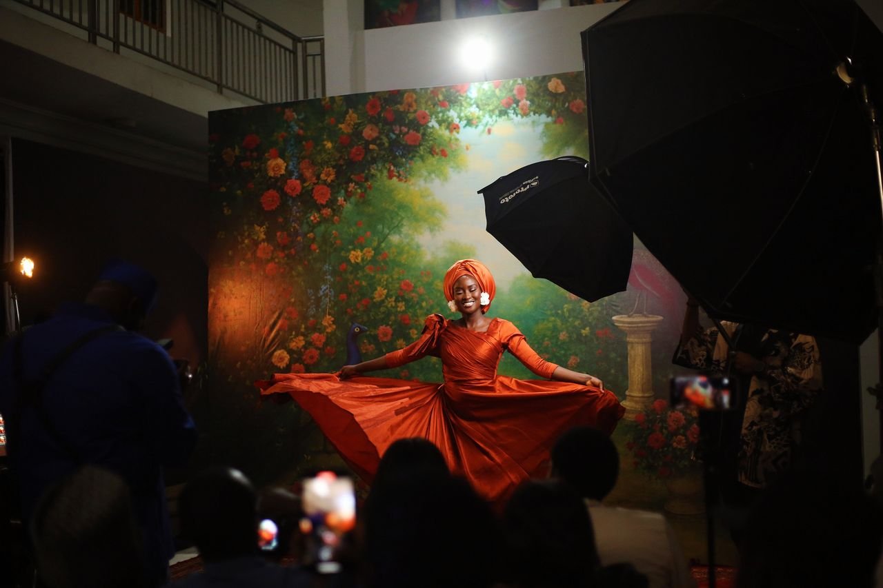 A woman in an orange dress poses gracefully indoors with a vibrant floral backdrop in Abuja, Nigeria.